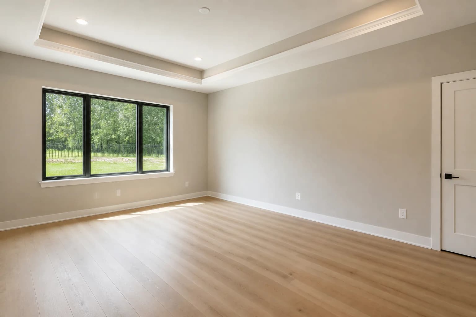 Completed empty new-construction primary bedroom with a tray ceiling, black-framed window, and light oak flooring