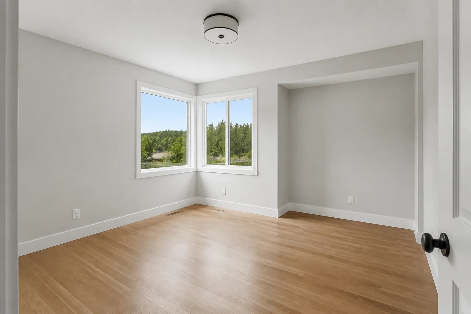 Vacant bonus room photographed with corner windows, a right-side alcove, gray walls, and oak flooring